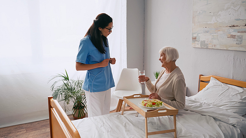 brunette nurse talking with happy aged patient near breakfast tray on bed