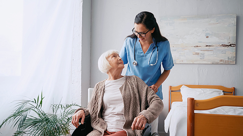 happy nurse in eyeglasses moving aged woman in wheelchair