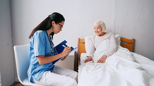 brunette nurse in eyeglasses writing prescription in clipboard near aged woman in bed