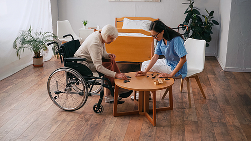 senior woman in wheelchair playing dominoes with brunette nurse in glasses