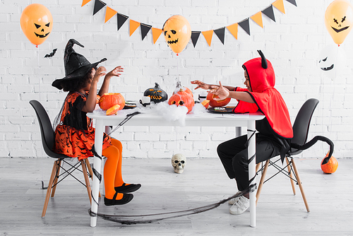 spooky african american kids in halloween costumes sitting at table with pumpkins and decoration during party