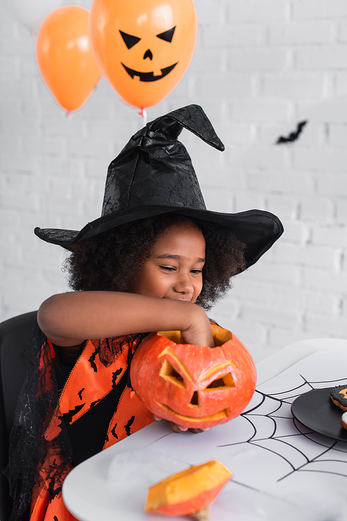 cheerful african american girl in witch halloween costume holding caved pumpkin