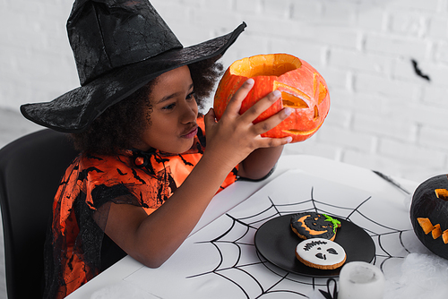 happy african american girl in witch halloween costume holding caved pumpkin