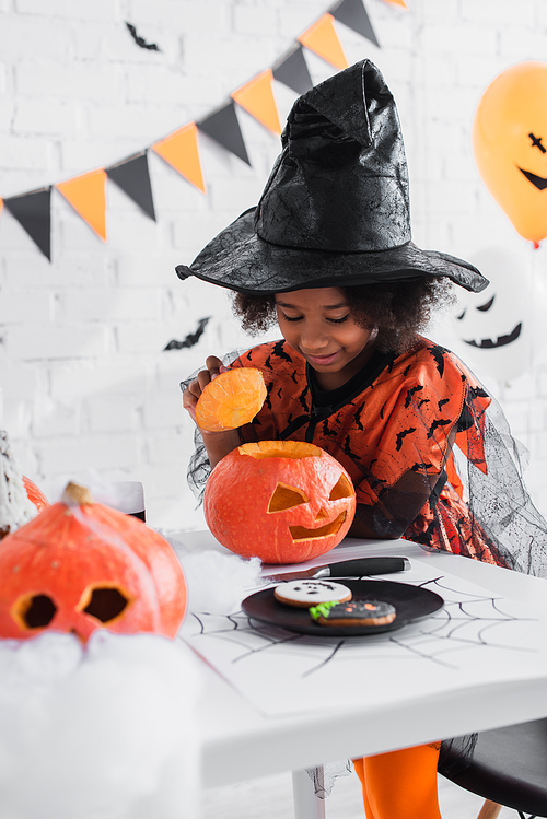 happy african american girl in witch halloween costume looking at caved pumpkin