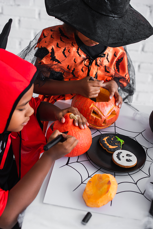 african american kids in halloween costumes carving and drawing on pumpkins near cookies