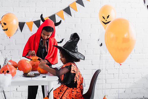 smiling african american kids in halloween costumes carving pumpkins near homemade cookies on table