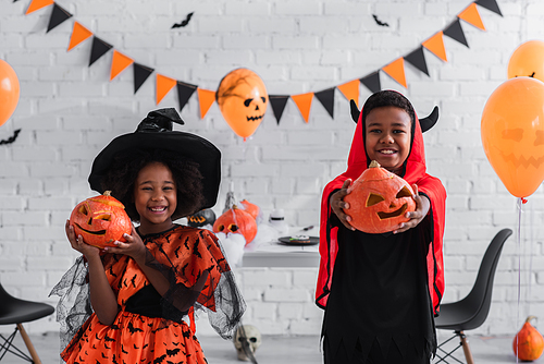 cheerful african american kids in halloween costumes holding carved pumpkins at home