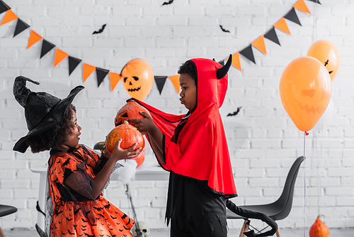side view of african american kids in halloween costumes holding carved pumpkins at home