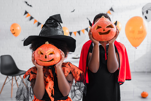 african american kids in halloween costumes covering faces with carved pumpkins at home