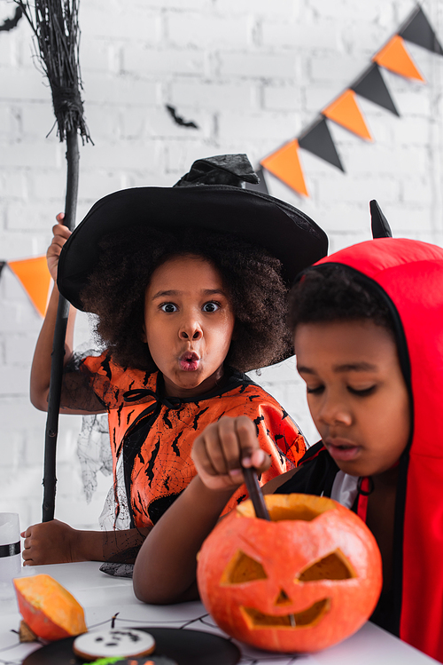 blurred african american boy in halloween costume carving pumpkin near spooky sister with broom