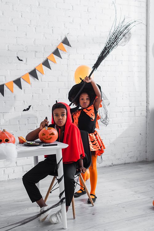african american boy in halloween costume carving pumpkin near spooky sister with broom