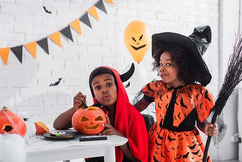 spooky african american boy in halloween costume carving pumpkin near sister with broom