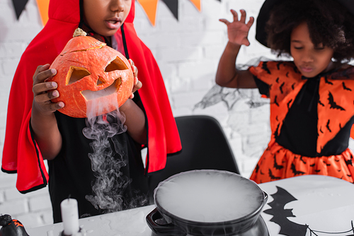 african american boy in halloween costume holding carved pumpkin with smoke while preparing potion near blurred sister