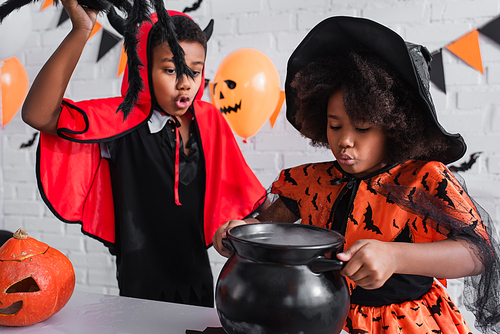 african american girl in halloween costume holding witch cauldron with potion near brother with toy spider