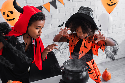 african american girl in halloween costume gesturing near witch cauldron with potion and brother with toy spider