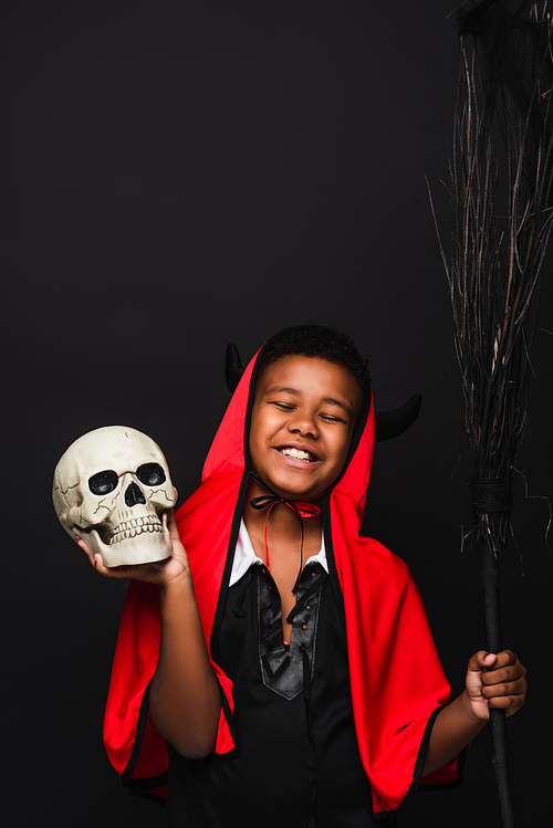 happy african american boy holding broom and skull isolated on black