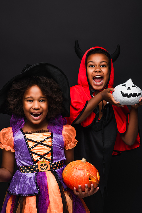 cheerful african american children in halloween costumes holding spooky pumpkins isolated on black
