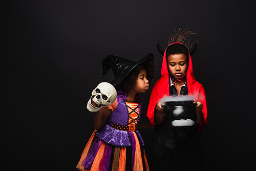 african american girl in halloween costume holding skull and blowing on witch cauldron with potion near brother isolated on black