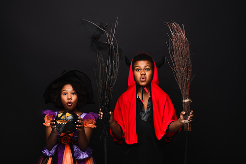 african american girl in halloween costume holding carved pumpkins near brother with brooms isolated on black