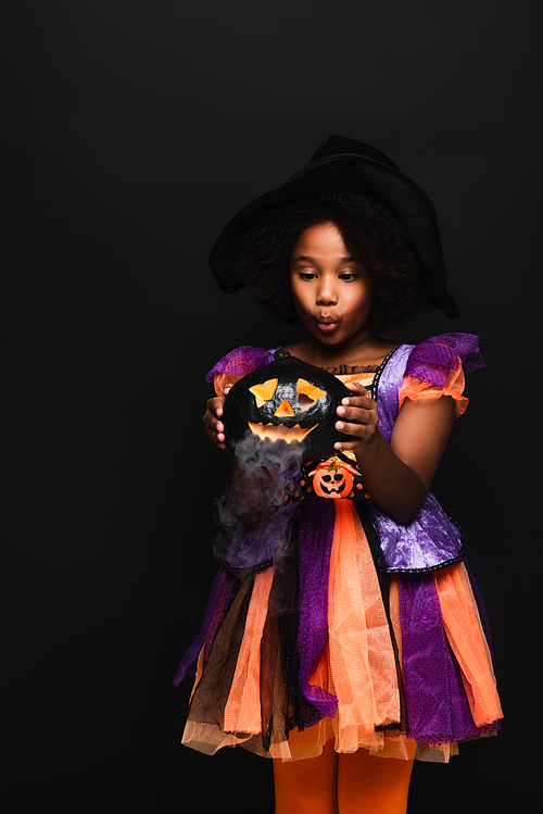 african american girl in halloween costume looking at carved pumpkin with smoke isolated on black