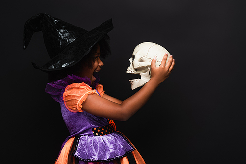 side view of excited african american girl in witch halloween costume holding skull isolated on black