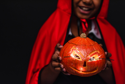 cropped view of blurred and happy african american boy in devil halloween costume holding carved pumpkin isolated on black