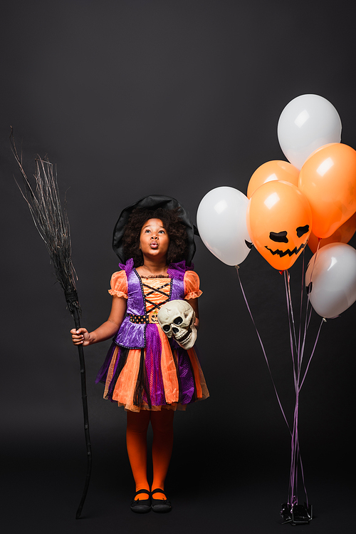 surprised african american girl in halloween costume holding broom and skull near balloons isolated on black