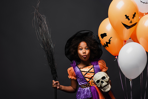 african american girl in halloween witch costume holding broom and skull near balloons isolated on black