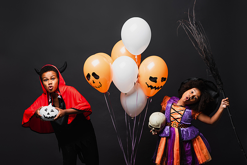 african american children in halloween costumes holding skull and pumpkin near balloons isolated on black