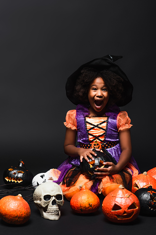 amazed african american girl in witch costume and pointed hat near carved pumpkins on black