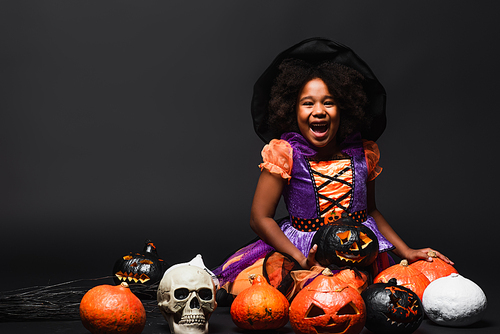 excited african american girl in witch costume and pointed hat near carved pumpkins on black