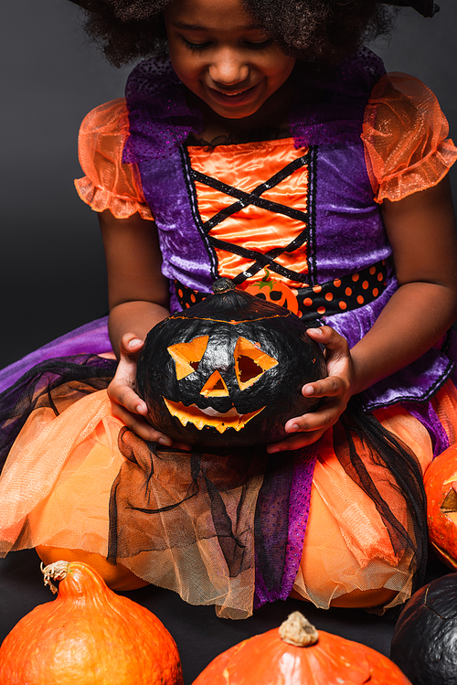 cheerful african american girl in costume holding carved pumpkin isolated on black