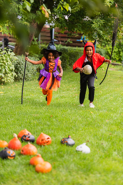excited african american children in halloween costumes holding brooms and running on lawn