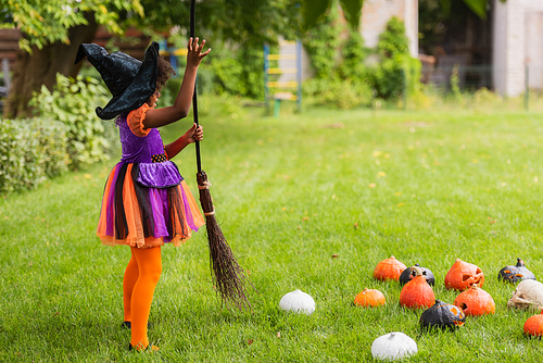 african american kid in witch halloween costume holding broom near carved pumpkins on lawn