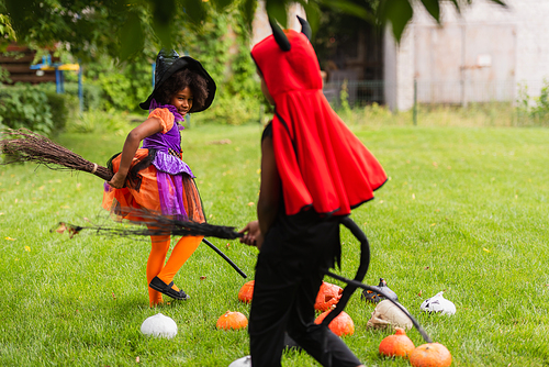 african american siblings in halloween costumes holding brooms and playing on lawn