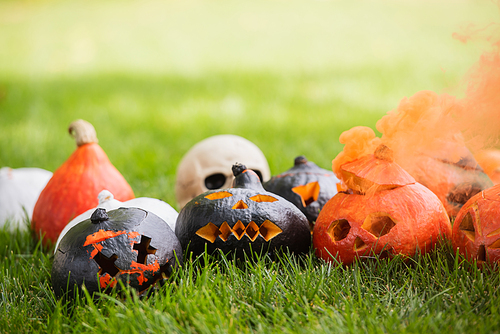 carved and scary pumpkins with orange smoke near blurred skull on green lawn