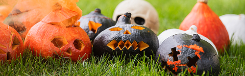 carved and scary pumpkins with orange smoke near blurred skull on green lawn, banner