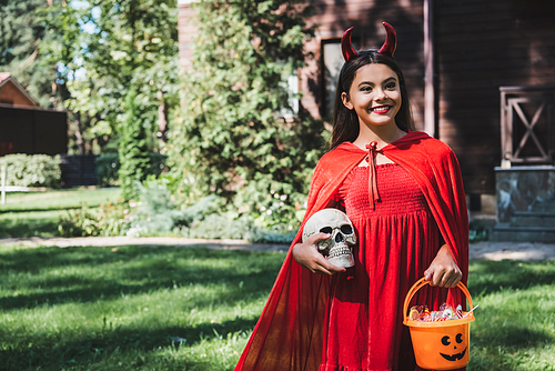 happy child in red demon costume standing with skull and bucket of candies outdoors