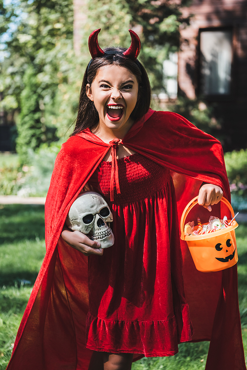 thrilled girl in devil halloween costume screaming while holding skull and bucket of candies