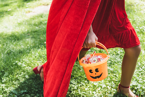 cropped view of girl in red costume carrying halloween bucket with candies while walking on grass