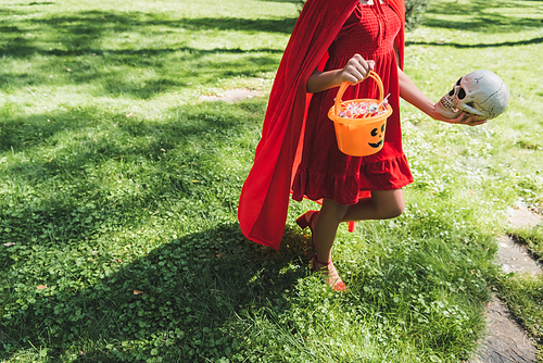 cropped view of child in red halloween costume walking on lawn with skull and bucket of sweets