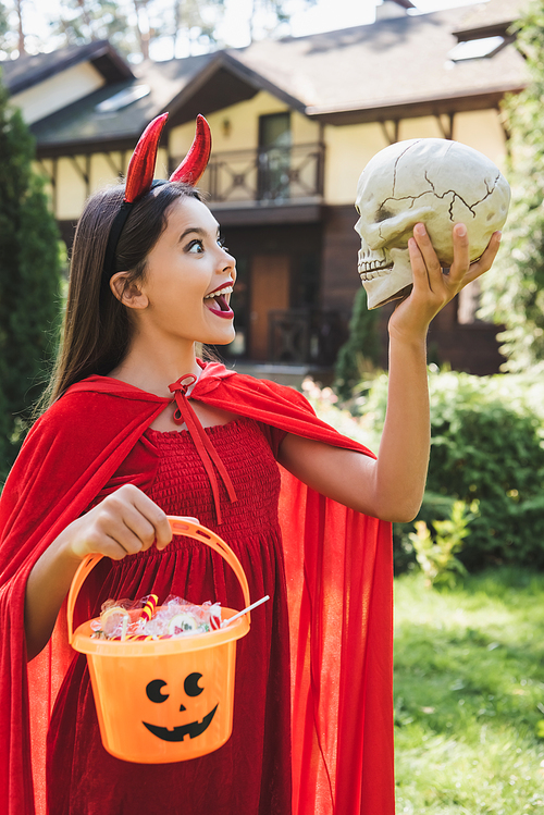 amazed girl in devil halloween costume looking at spooky skull while holding bucket with candies