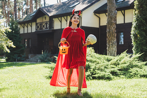 joyful girl in demon halloween costume holding skull and bucket with candies near blurred house