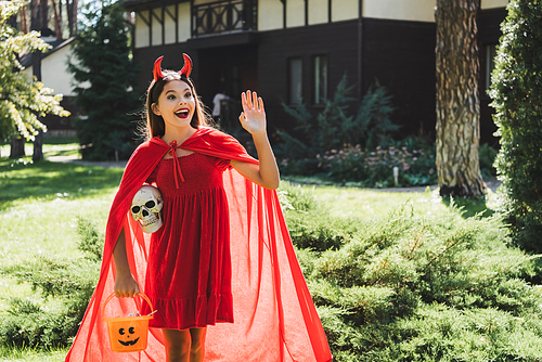 amazed girl in devil halloween costume waving hand while standing with skull and bucket of candies near blurred house