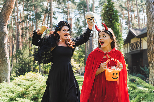 girl in devil halloween costume holding bucket with candies and screaming near mom with skull and toy hand