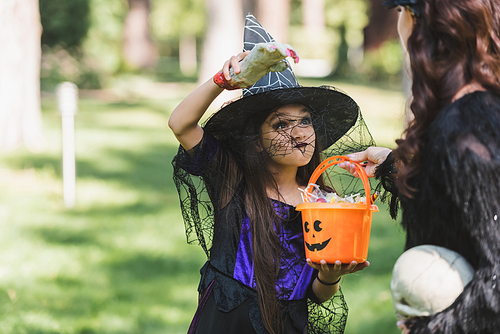 girl in witch hat holding bucket with candies and scaring mom with toy hand