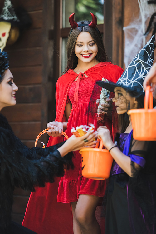 mom giving candies to happy daughters in devil and witch halloween costumes