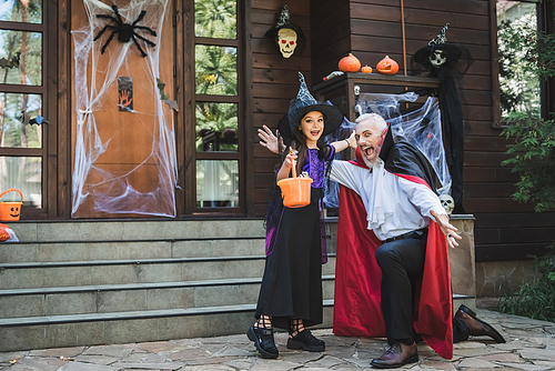 excited man in vampire costume and girl with bucket of sweets near porch with halloween decoration