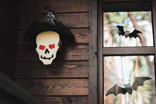 paper cut skull in witch hat near black bats on window of wooden cottage