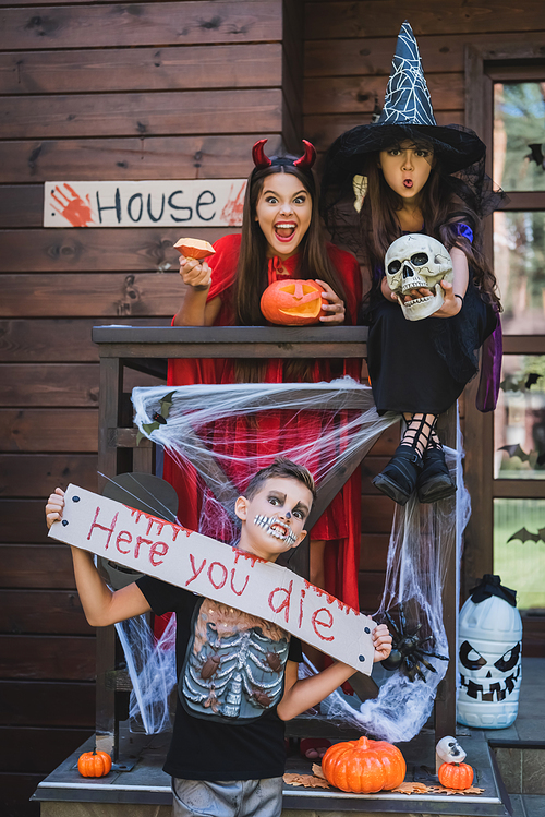 boy with scary grimace holding card with here you die lettering near spooky sisters in halloween costumes